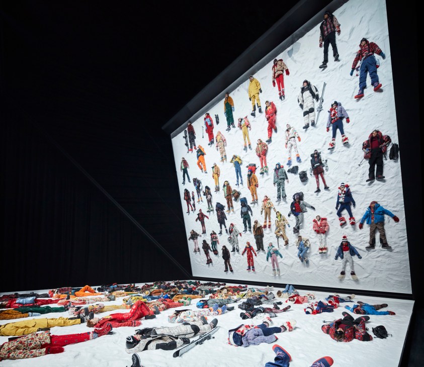 Art installation showing people in colorful ski gear on snowy slope, with similar figures scattered on floor below