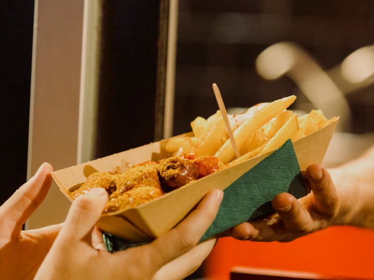 Hands holding paper container of currywurst with fries and curry sauce being prepared fresh