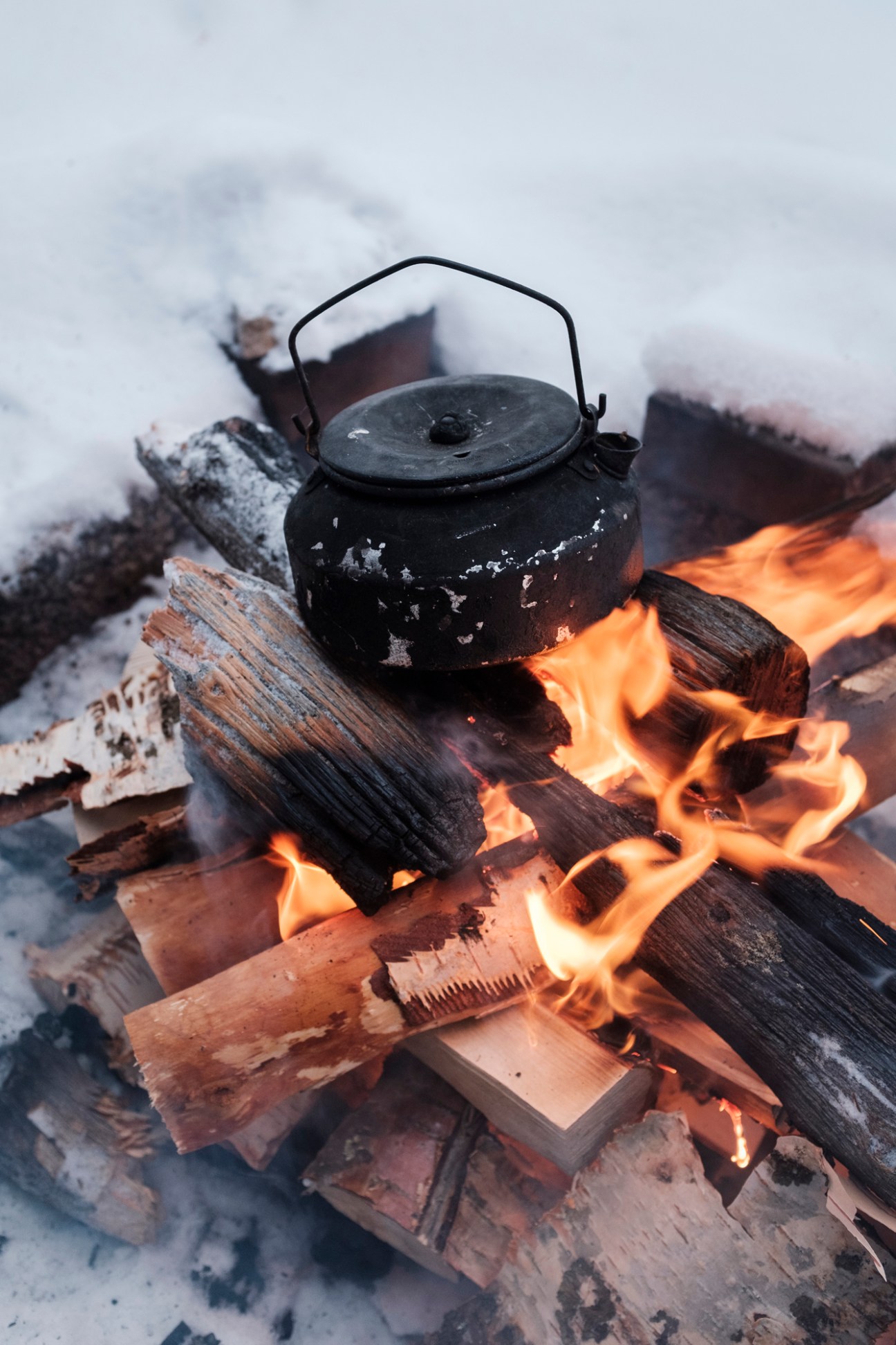 Black kettle heating over campfire with burning logs and snow in background