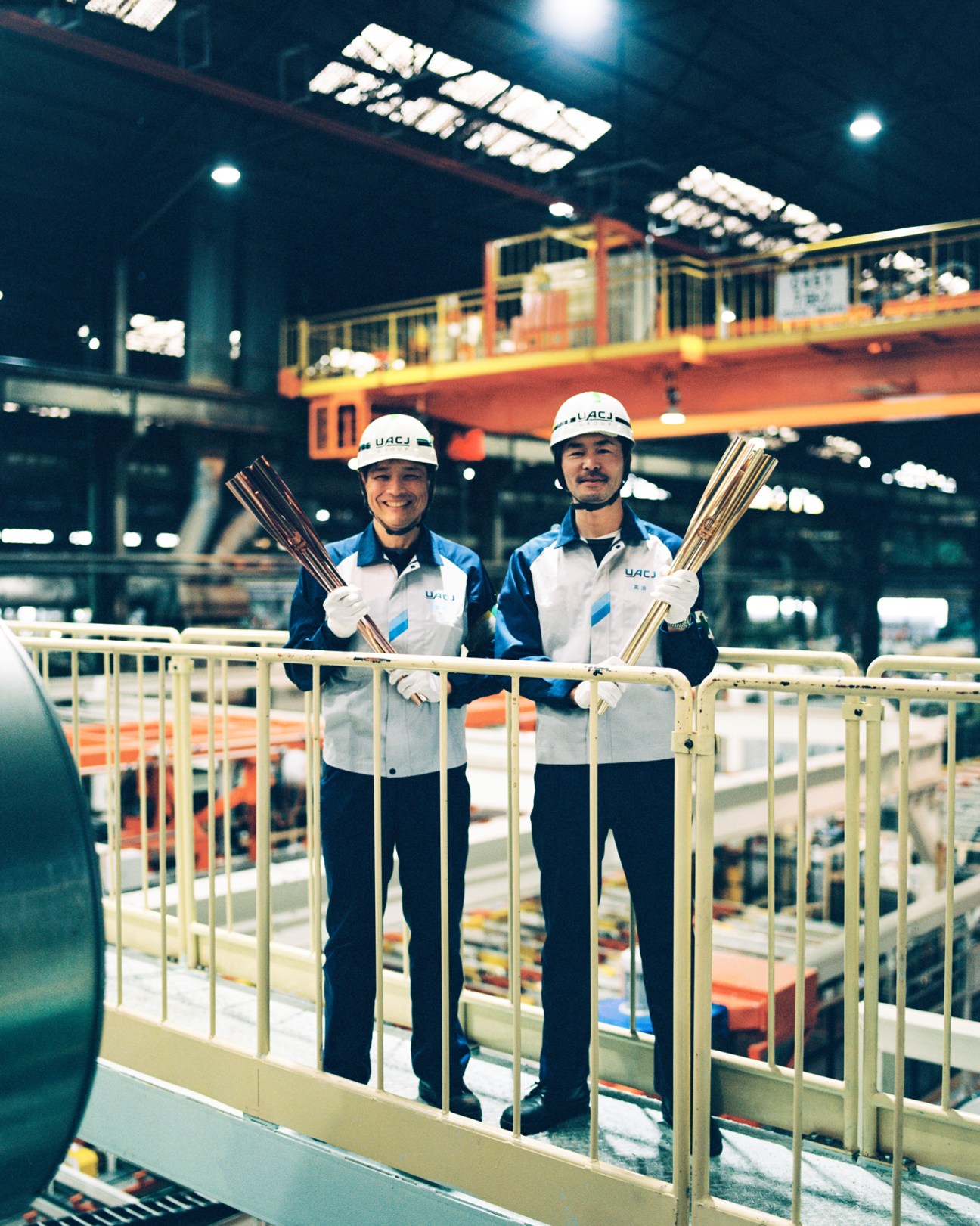 Two workers in white hard hats and UACJ branded uniforms holding metallic torch-like objects in an industrial facility