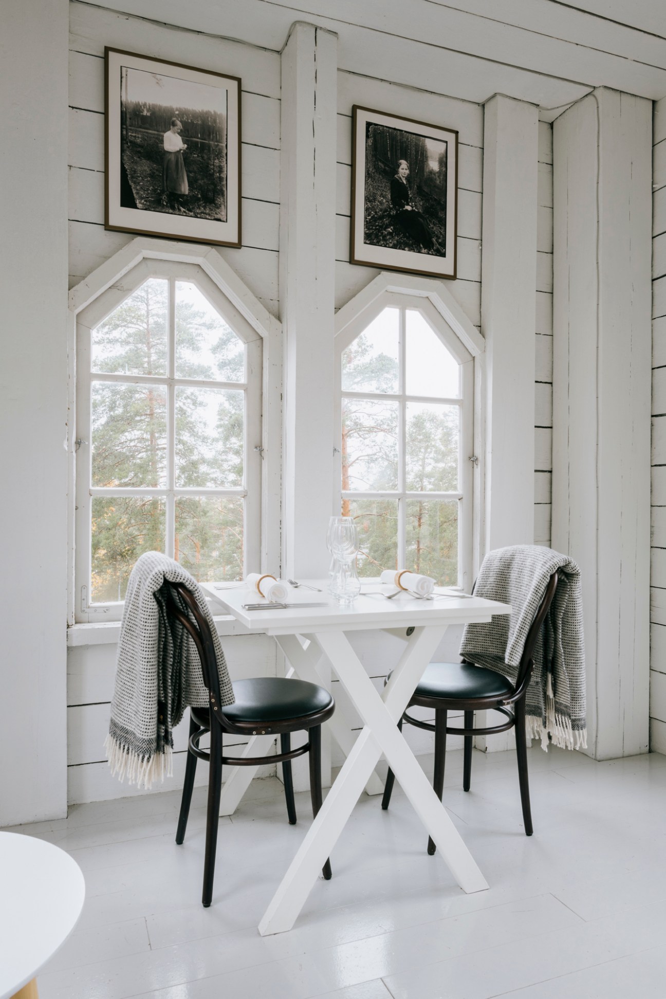 Minimalist dining setup with white table, black chairs, and framed black-and-white photographs above large arched windows overlooking forest.