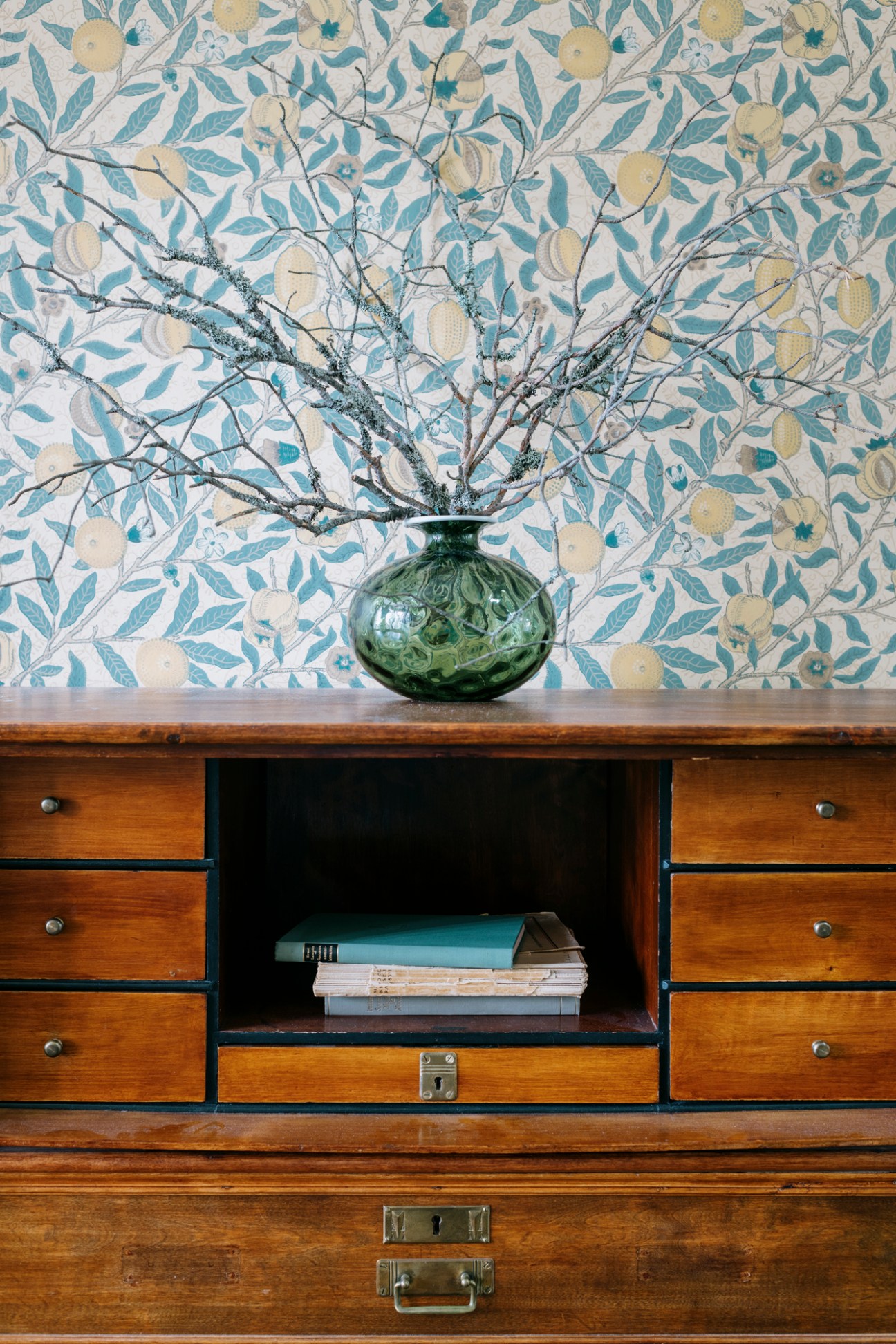 Green glass vase with bare branches against blue and cream botanical wallpaper on wooden dresser.