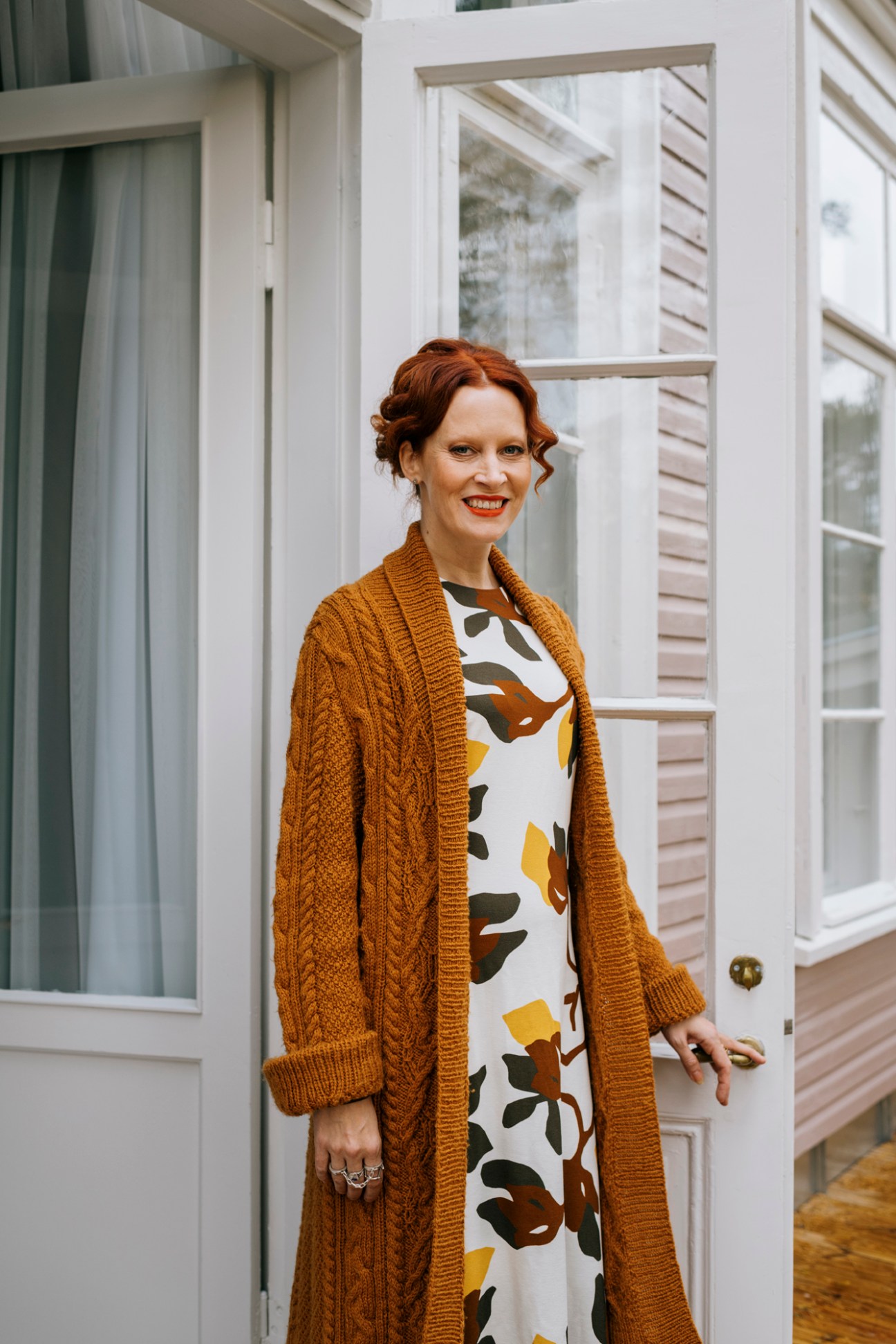 Woman with reddish hair wearing a mustard cardigan and patterned dress standing in a doorway with white paneled doors behind her.