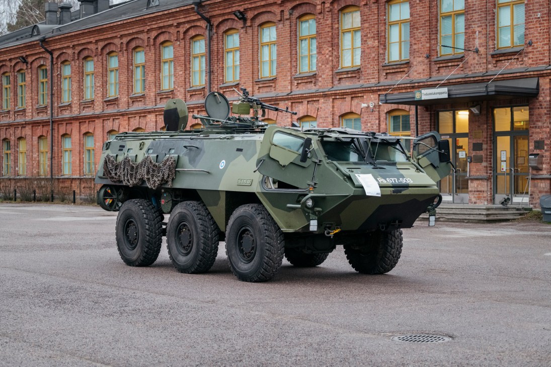 Green six-wheeled Pasi armoured personnel carrier parked on street in front of red brick building