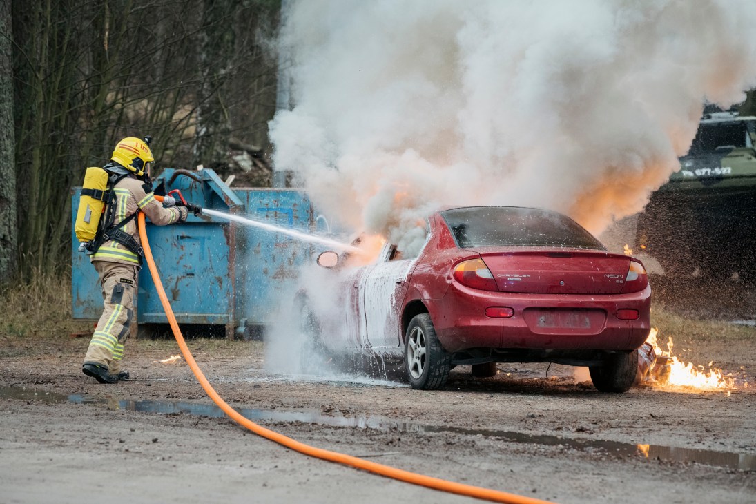 Firefighter in yellow helmet and protective gear spraying water on a burning red car with thick smoke