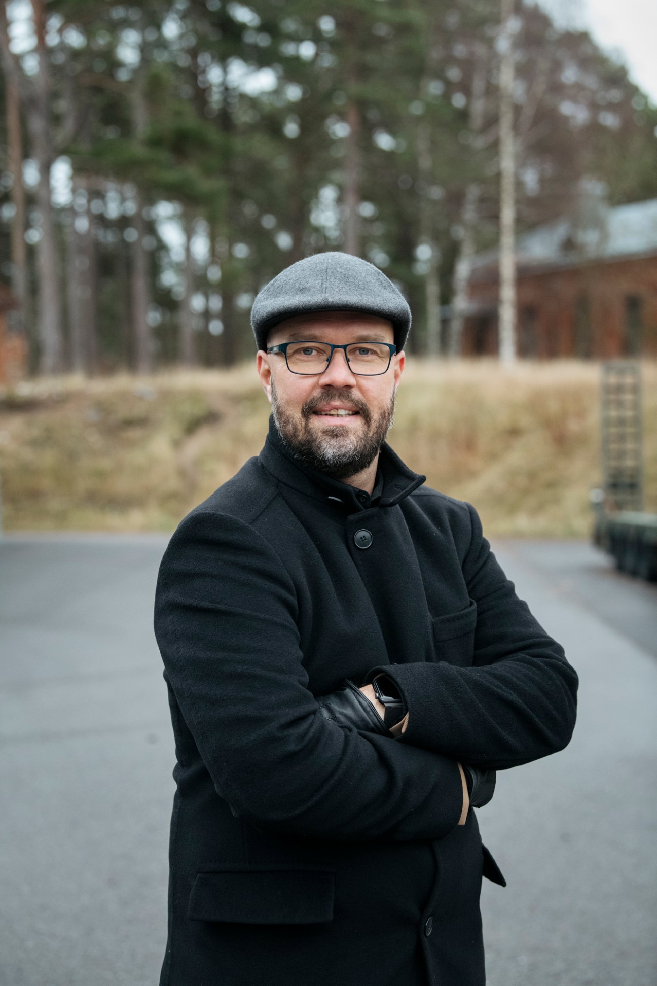 Man in grey flat cap, glasses, and dark coat with arms crossed standing outdoors