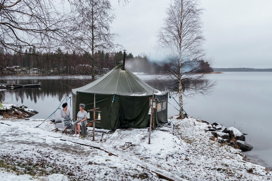 Two people in robes sitting outside a green military-style tent with chimney pipe by a snowy lakeside