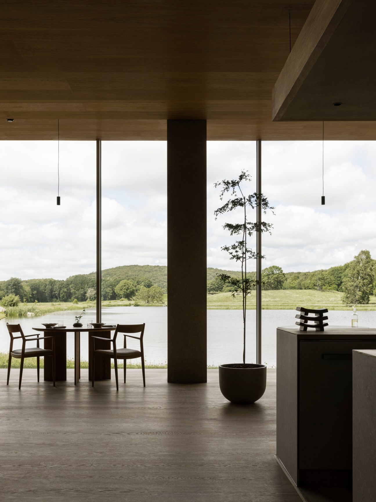 Modern restaurant interior with floor-to-ceiling windows overlooking a lake, featuring a dining table and indoor plant