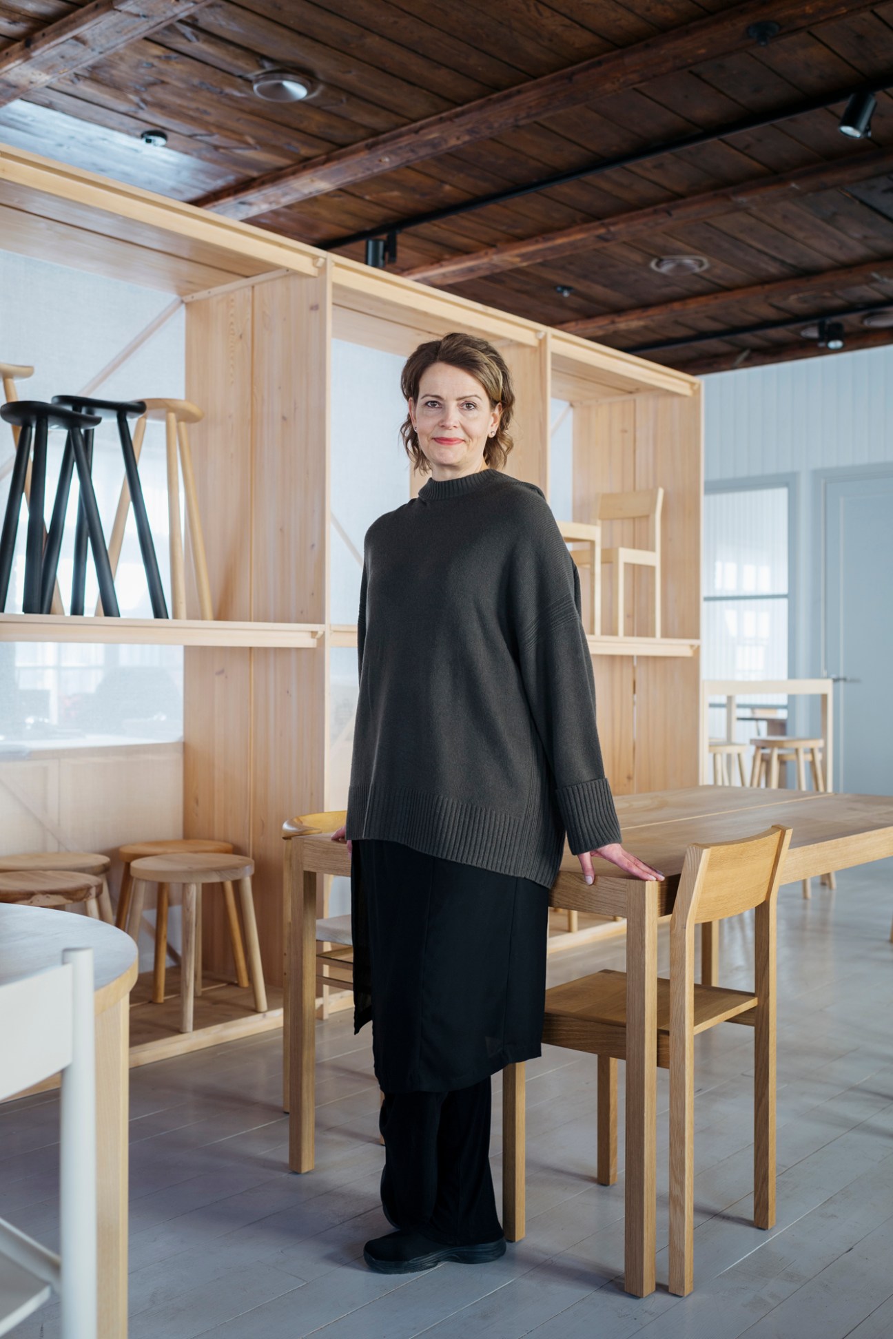 Woman in black sweater standing in wooden interior with light wood furniture and shelving