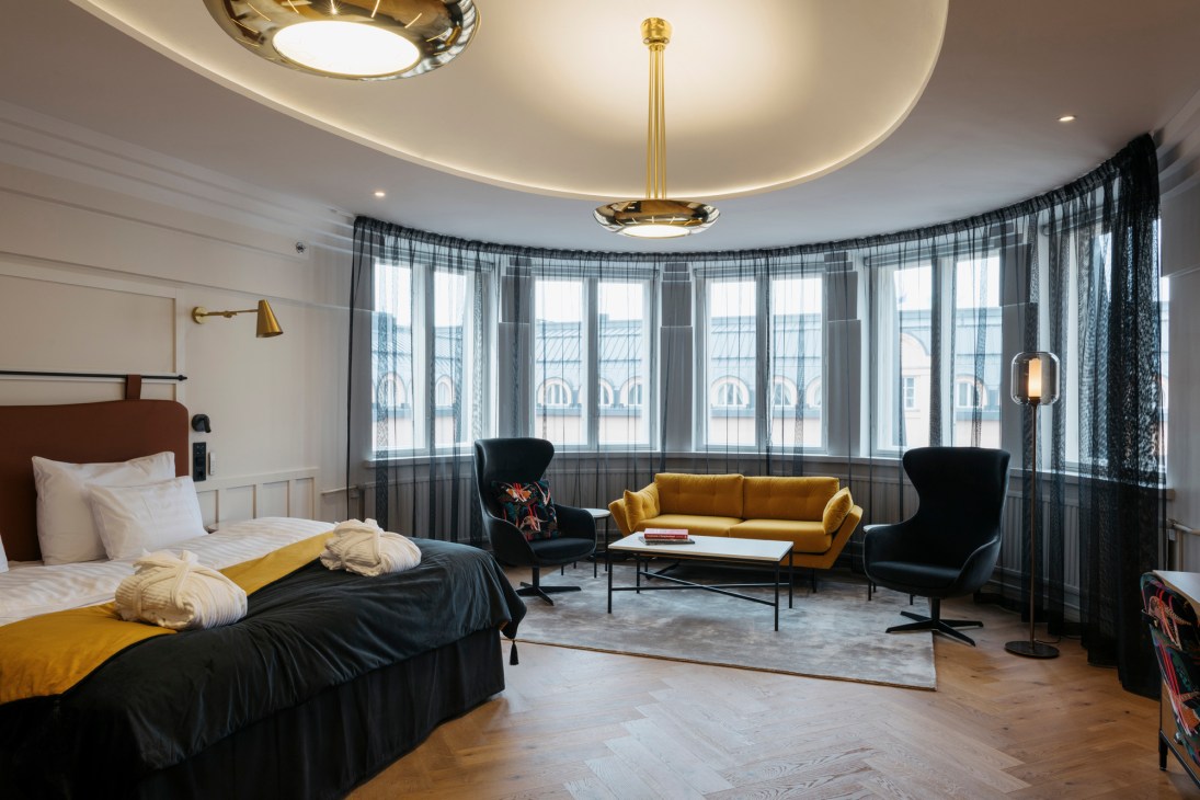 Junior suite interior with curved bay windows, yellow sofa, dark armchairs and ornate ceiling fixture