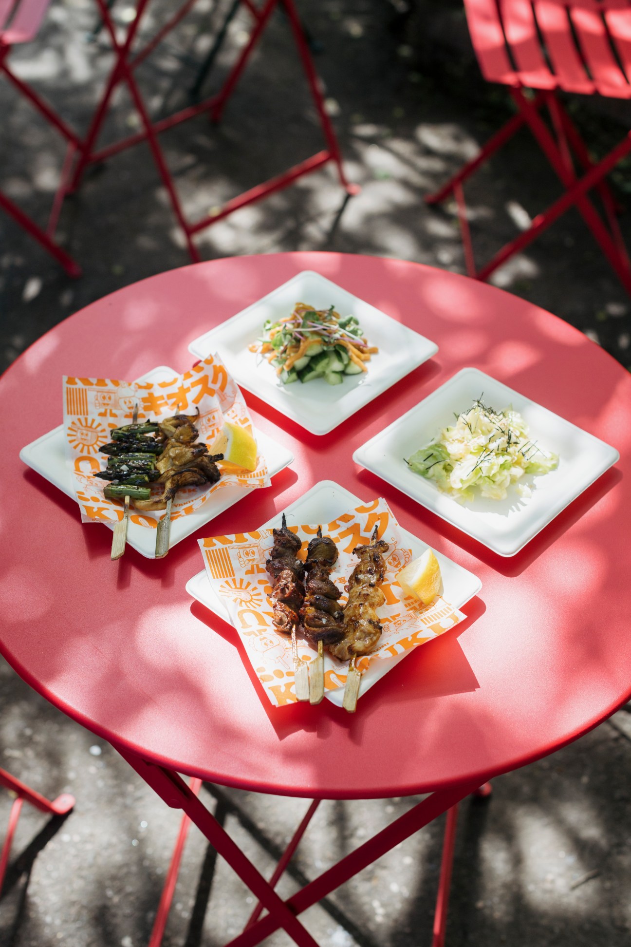 Yakitori skewers and vegetable dishes on white plates arranged on a pink table at Kiosuku.