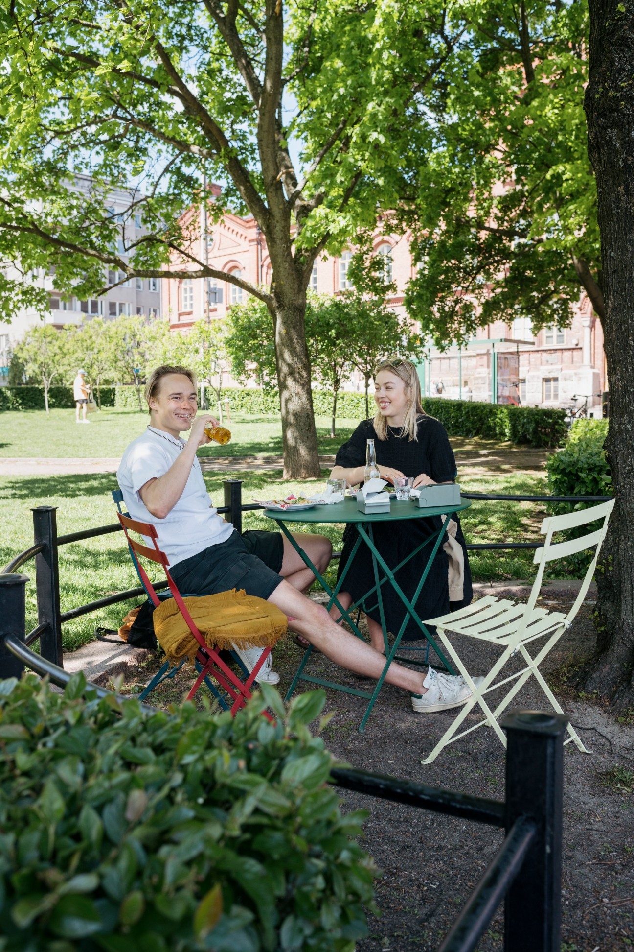 Two people dining outdoors at a green table in a Helsinki park, with historic buildings visible behind trees.