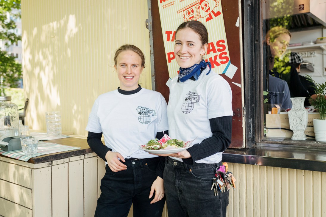 Two smiling staff members in matching white shirts with globe logos at Kiosquito kiosk window.