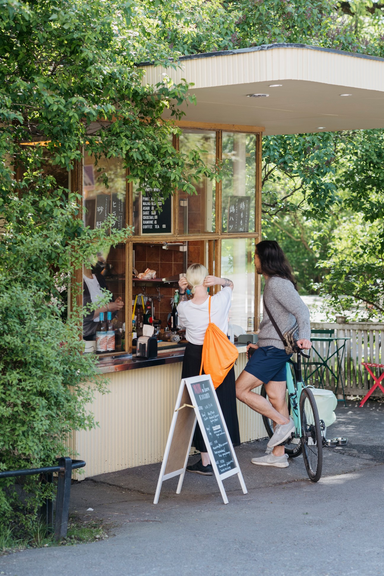 Customers order ice lollies at a Helsinki kiosk surrounded by green foliage.