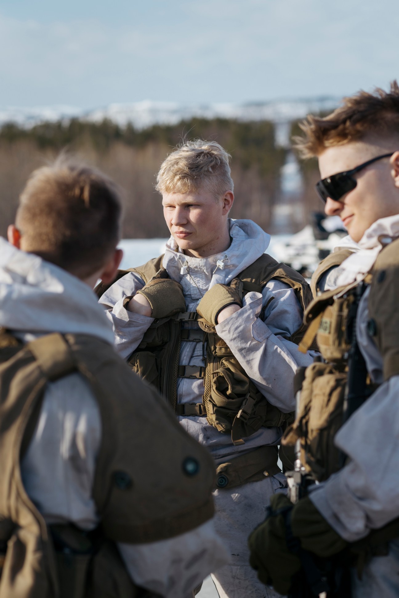 Norwegian soldier in white winter gear checking rifle sight on tripod against snowy landscape.