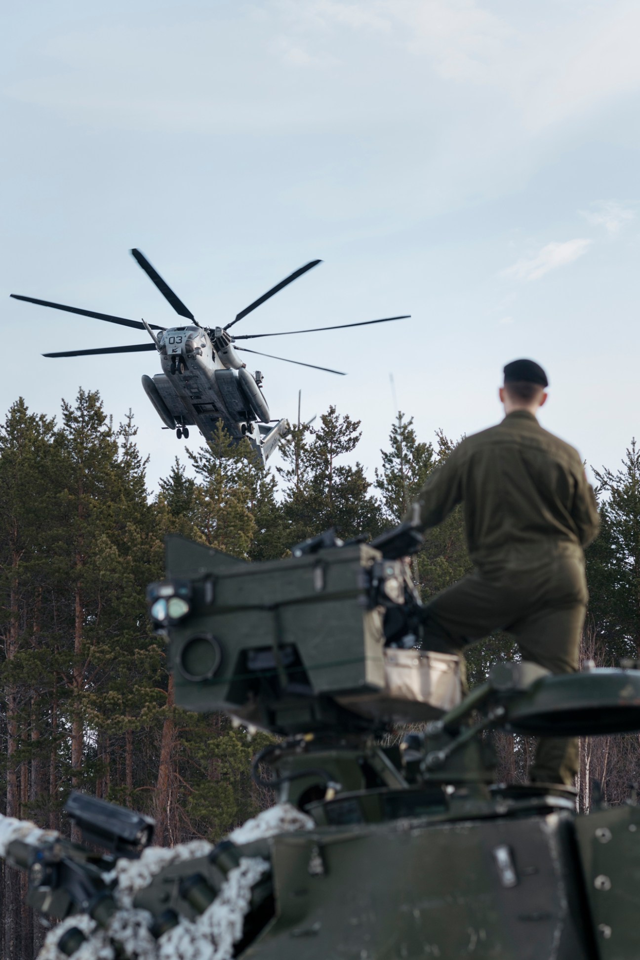 A Sikorsky CH-53E Super Stallion helicopter hovers above a snowy landscape with a soldier in olive fatigues watching from the ground.