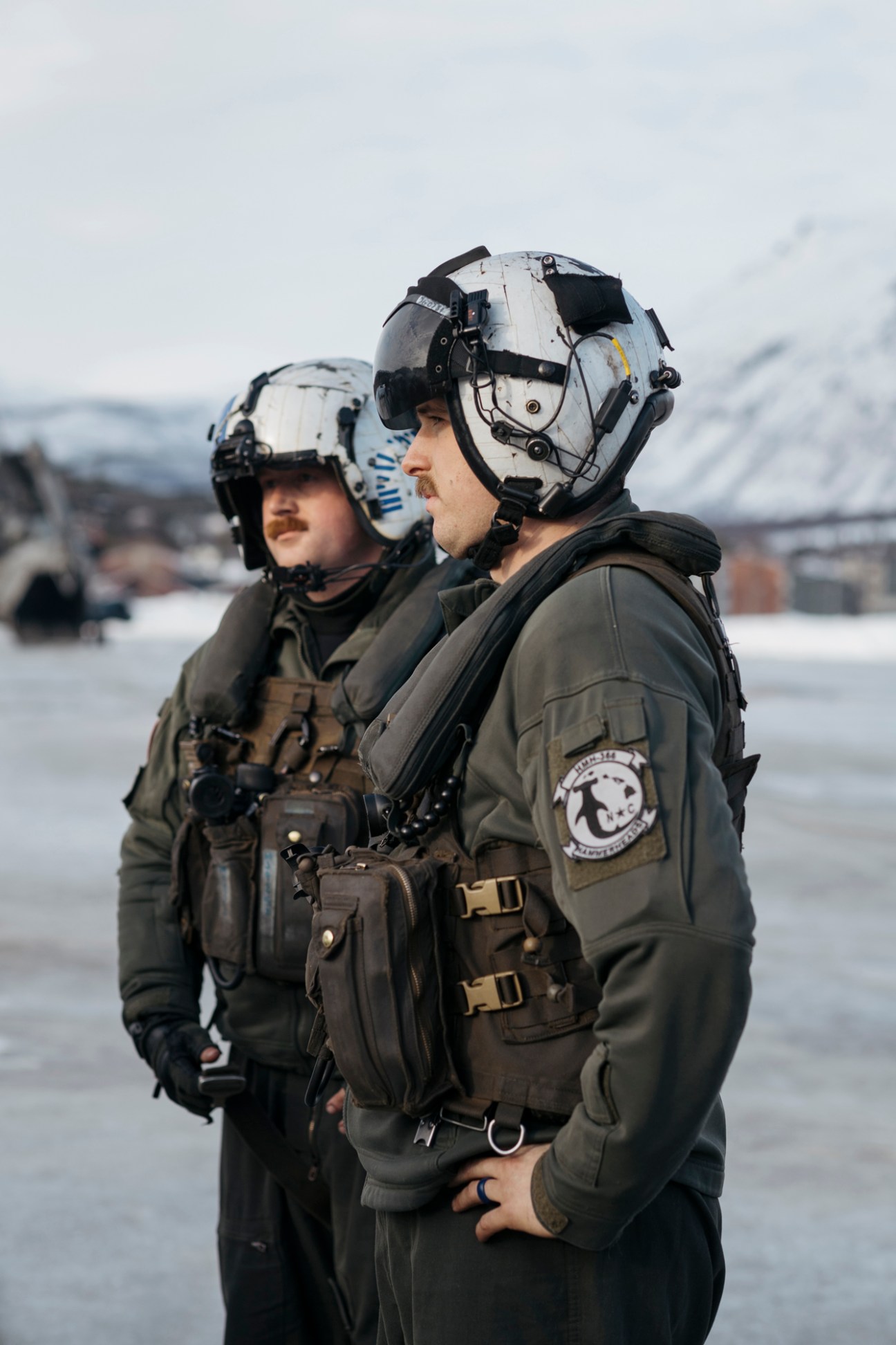Two US Marine Corps pilots in flight suits and white helmets stand on an icy field during a NATO training exercise.