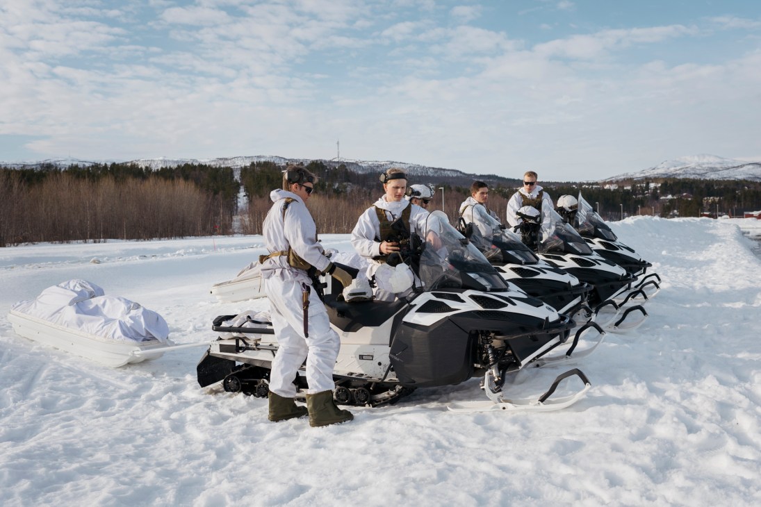 Norwegian military conscripts in winter white camouflage uniforms stand with snowmobiles on a snowy field during Cold Response exercise.