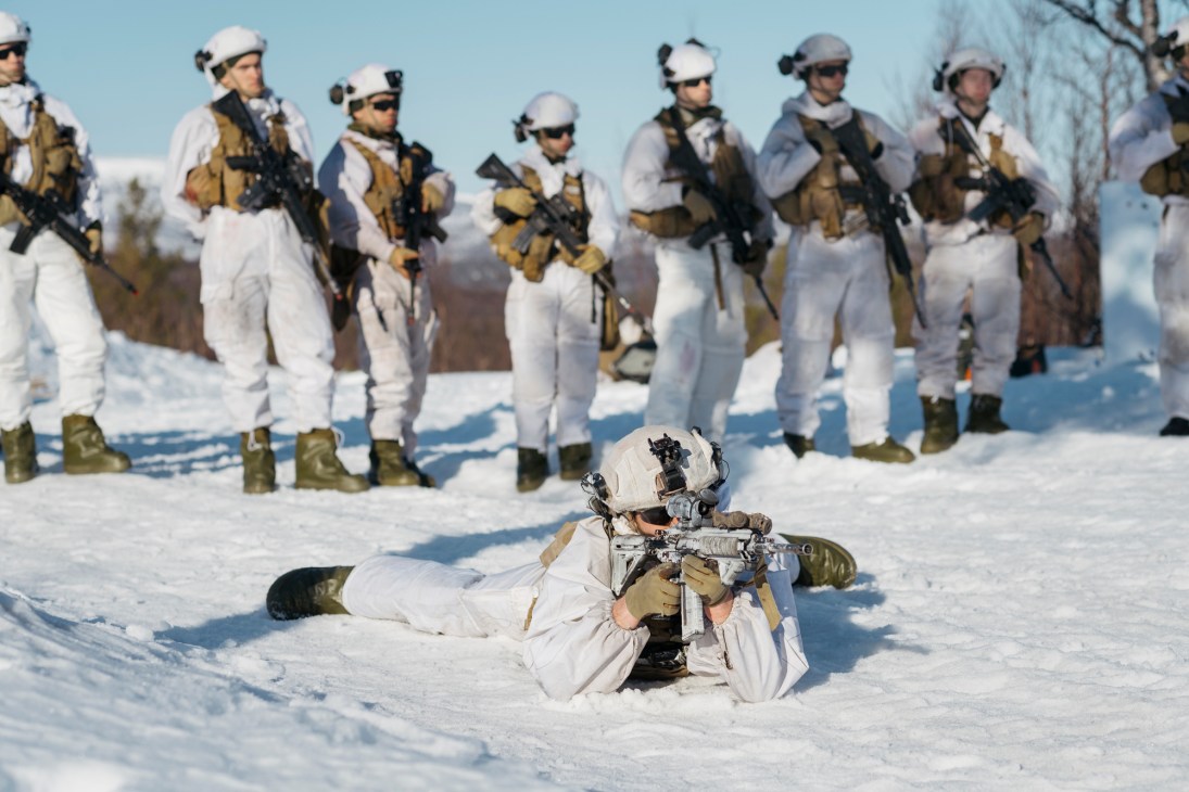 Norwegian conscript in white winter camouflage lying prone in snow with rifle while soldiers stand in formation behind.