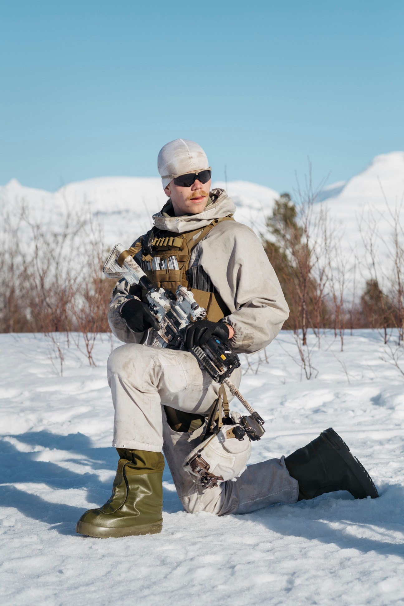 Armed soldier in white winter camouflage kneeling in snow with rifle and tactical gear.
