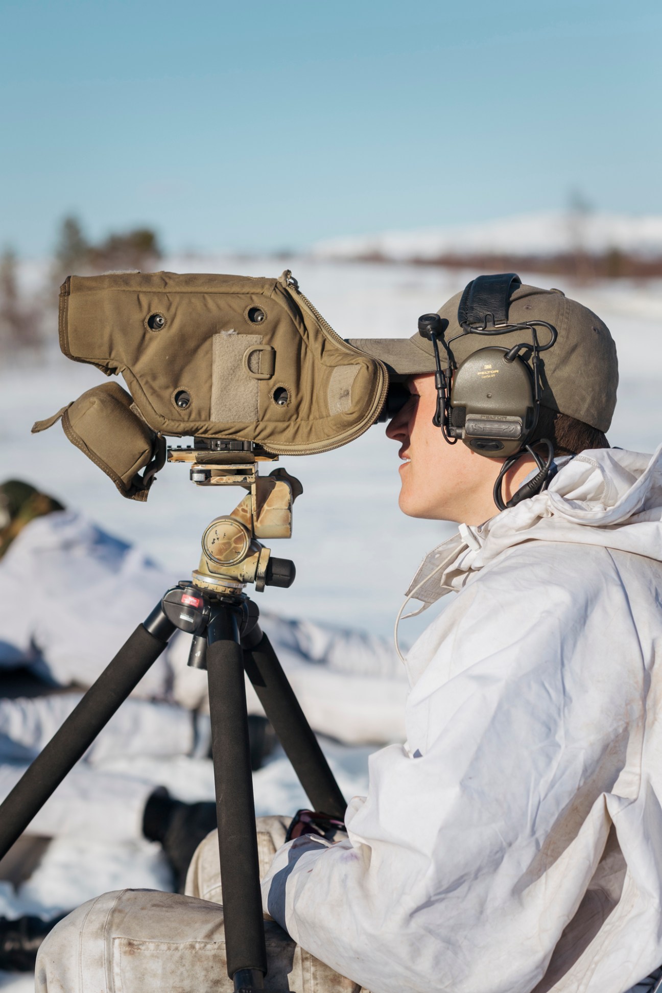 Norwegian soldier in white winter gear checking rifle sight on tripod against snowy landscape.