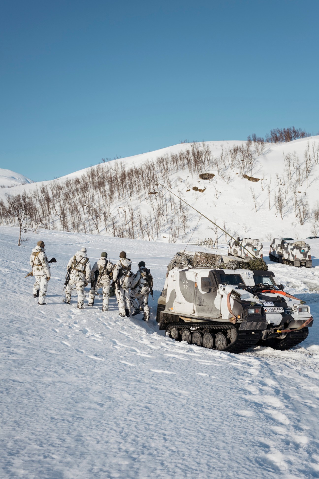 Norwegian military conscripts in winter white camouflage uniforms stand with snowmobiles on a snowy field during Cold Response exercise.