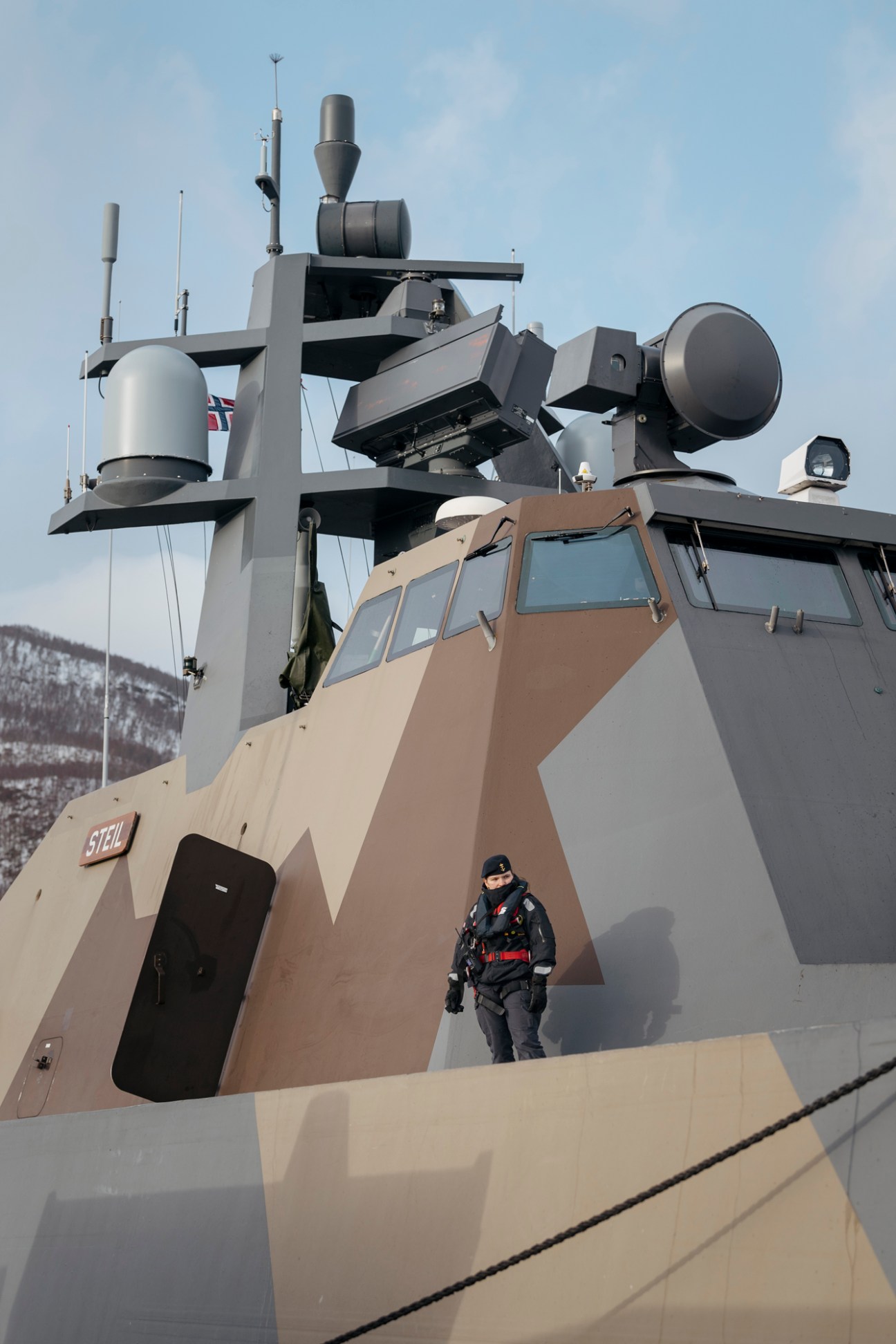 Norwegian navy's HNoMS Steil stealth corvette with angular hull and communications equipment, with a sailor standing on deck against snowy mountains.