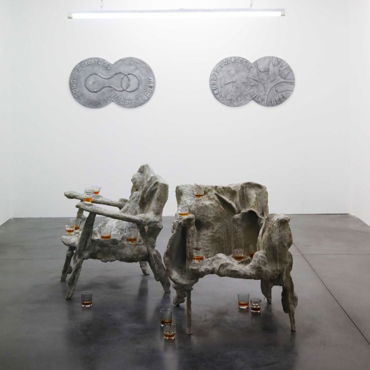 Two weathered stone chairs with whiskey glasses placed on and around them in a gallery installation