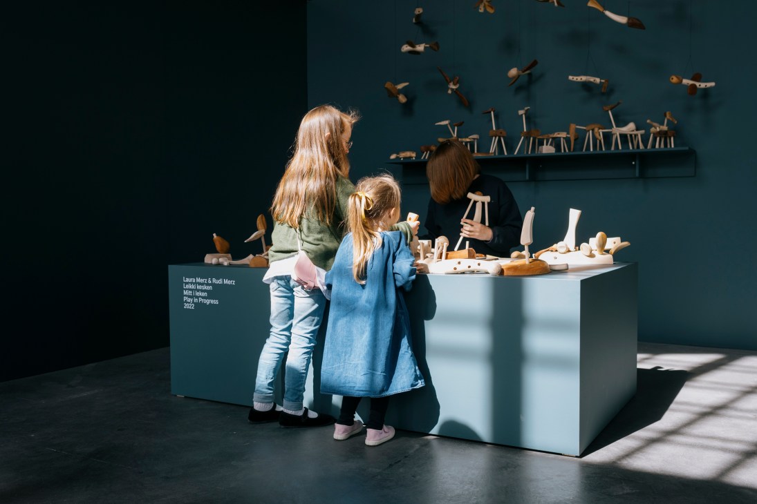 An adult and two children interact with a wooden sculptural installation at an exhibition, with wooden tools and objects displayed on a white table and mounted on a dark blue wall behind them.