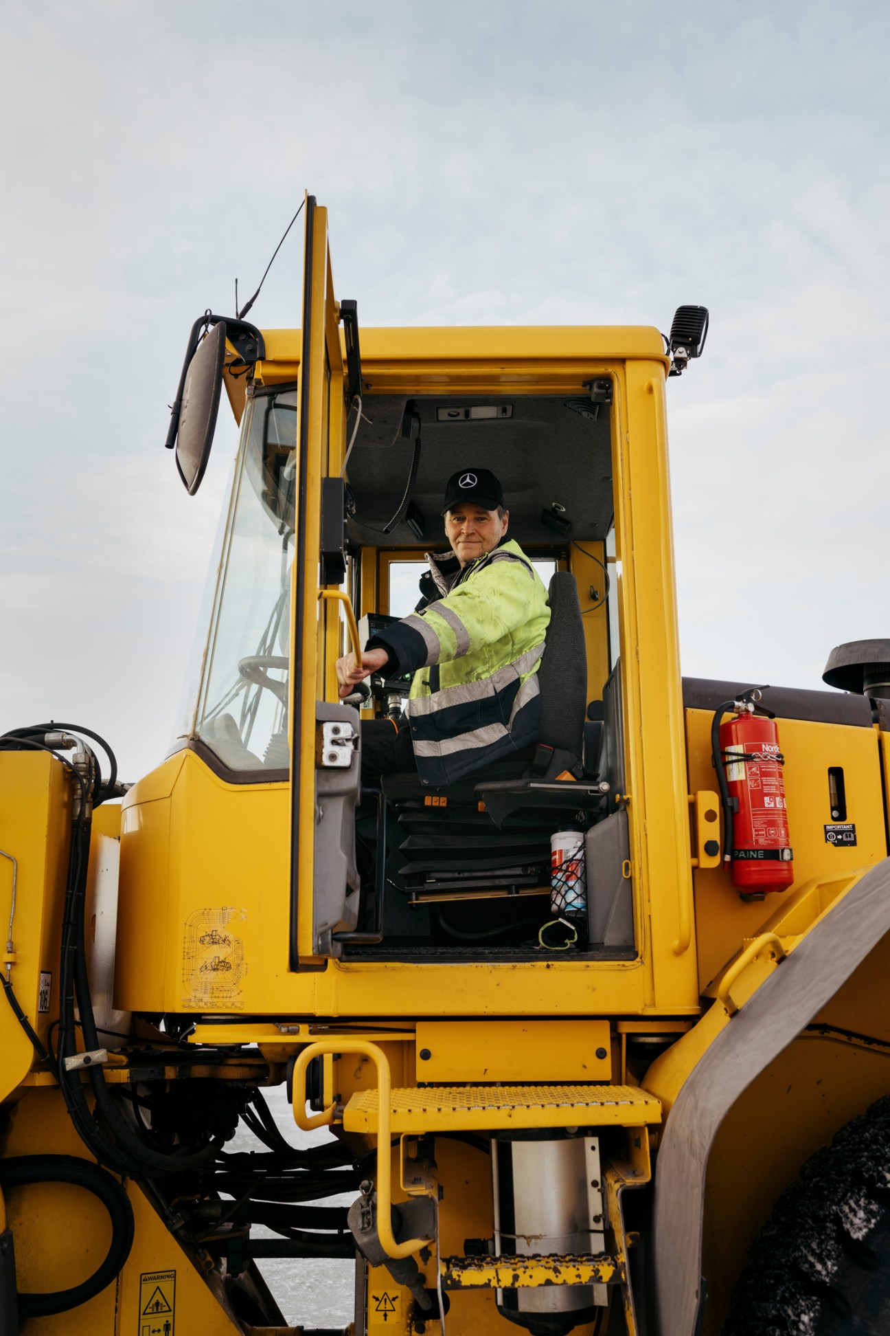 Man in high-visibility jacket and Mercedes cap sitting in yellow heavy machinery cab