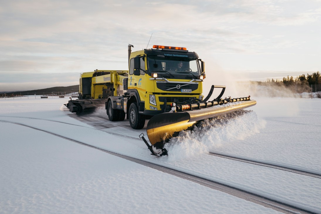 Yellow Volvo truck with large snow plow clearing snow from airport runway, throwing snow to the side
