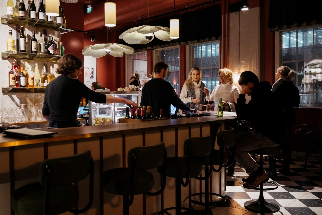 Interior view of a modern bar with pendant lights, shelves of bottles, bartenders serving customers at wooden counter