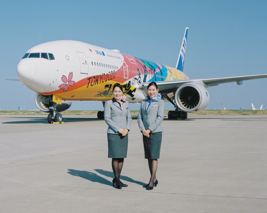 Two female flight attendants in grey uniforms standing in front of ANA's Olympic-themed Boeing 777 aircraft
