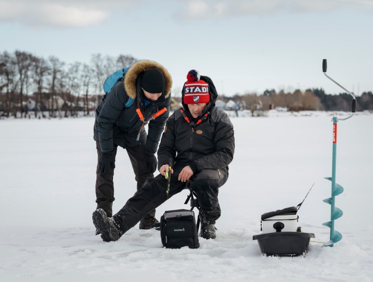 Two people ice fishing on frozen lake, crouched around equipment including fish finder and auger in snowy landscape