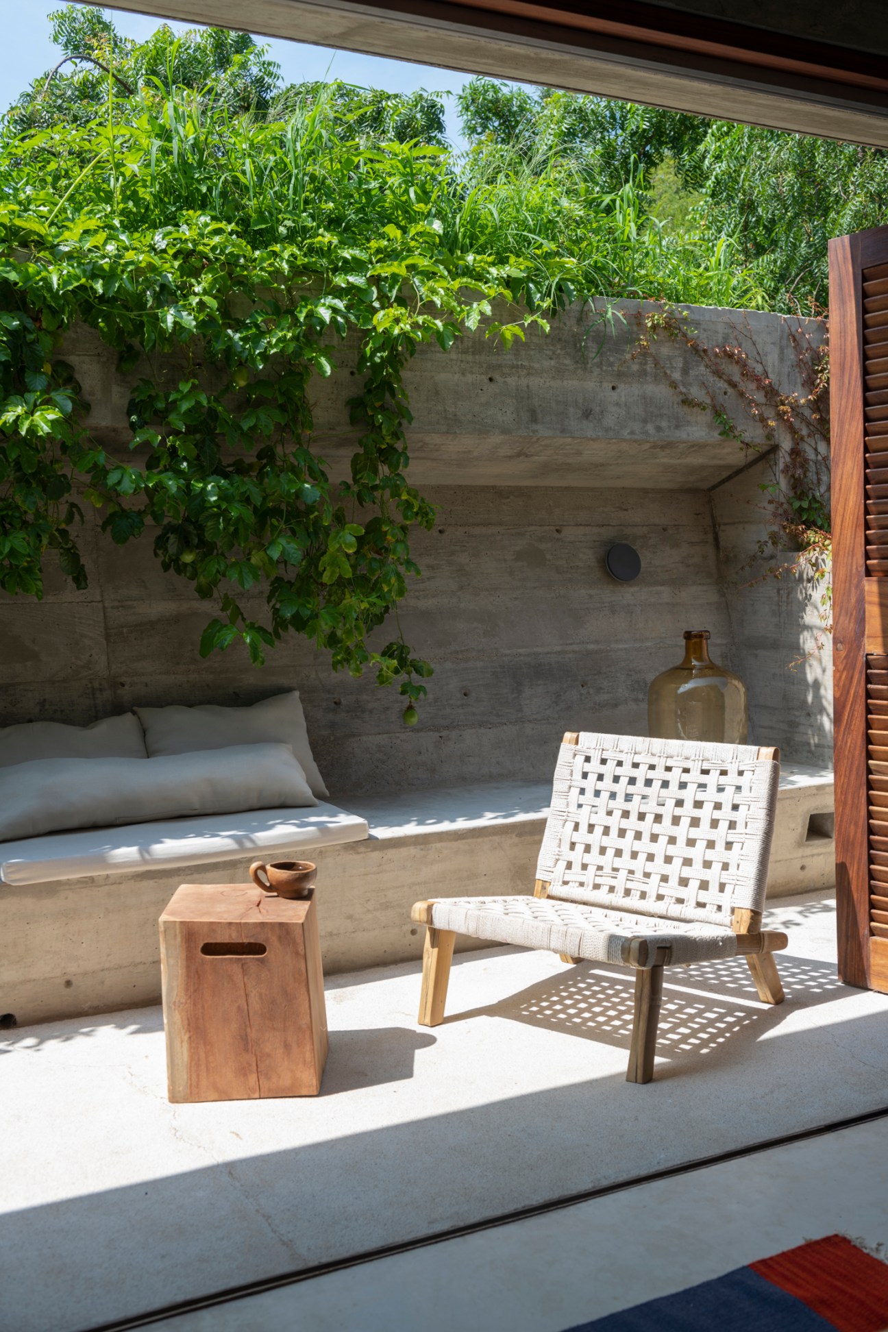 Woven chair and wooden side table on outdoor terrace with lush green vines growing on concrete wall
