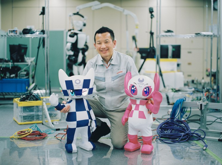 Man posing with the two Tokyo Olympics mascot robots in a factory.