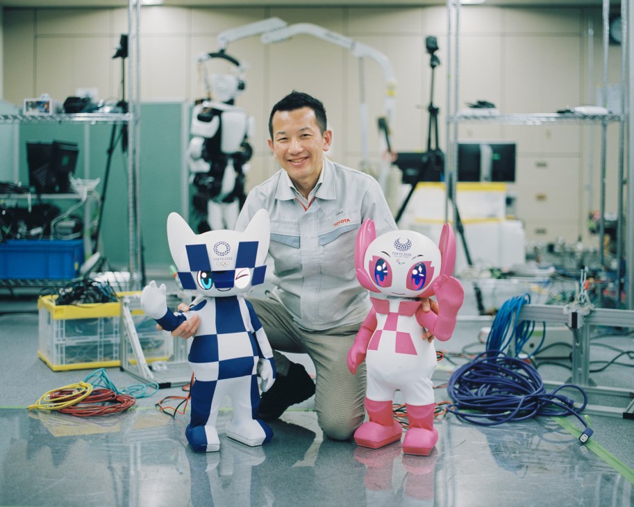 Tomohisa Moridaira kneeling in a laboratory between two robotic Olympic mascots Miraitowa and Someity