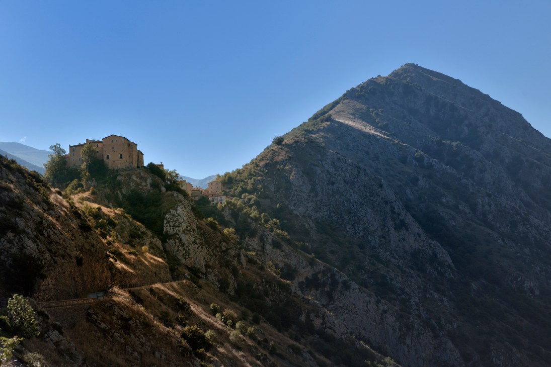 Medieval stone buildings perched on a rocky hillside with dramatic Apennine mountain peaks in the background under clear blue sky.
