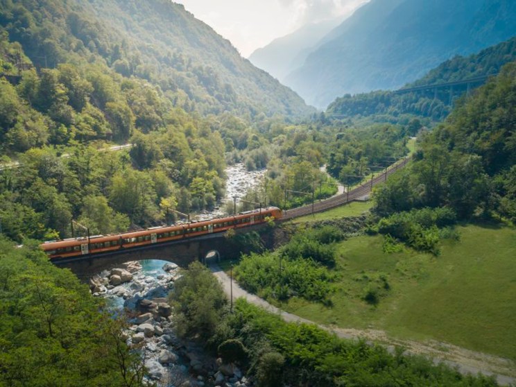 Orange Treno Gottardo train crosses a bridge over a river through a forested Alpine valley in Switzerland.
