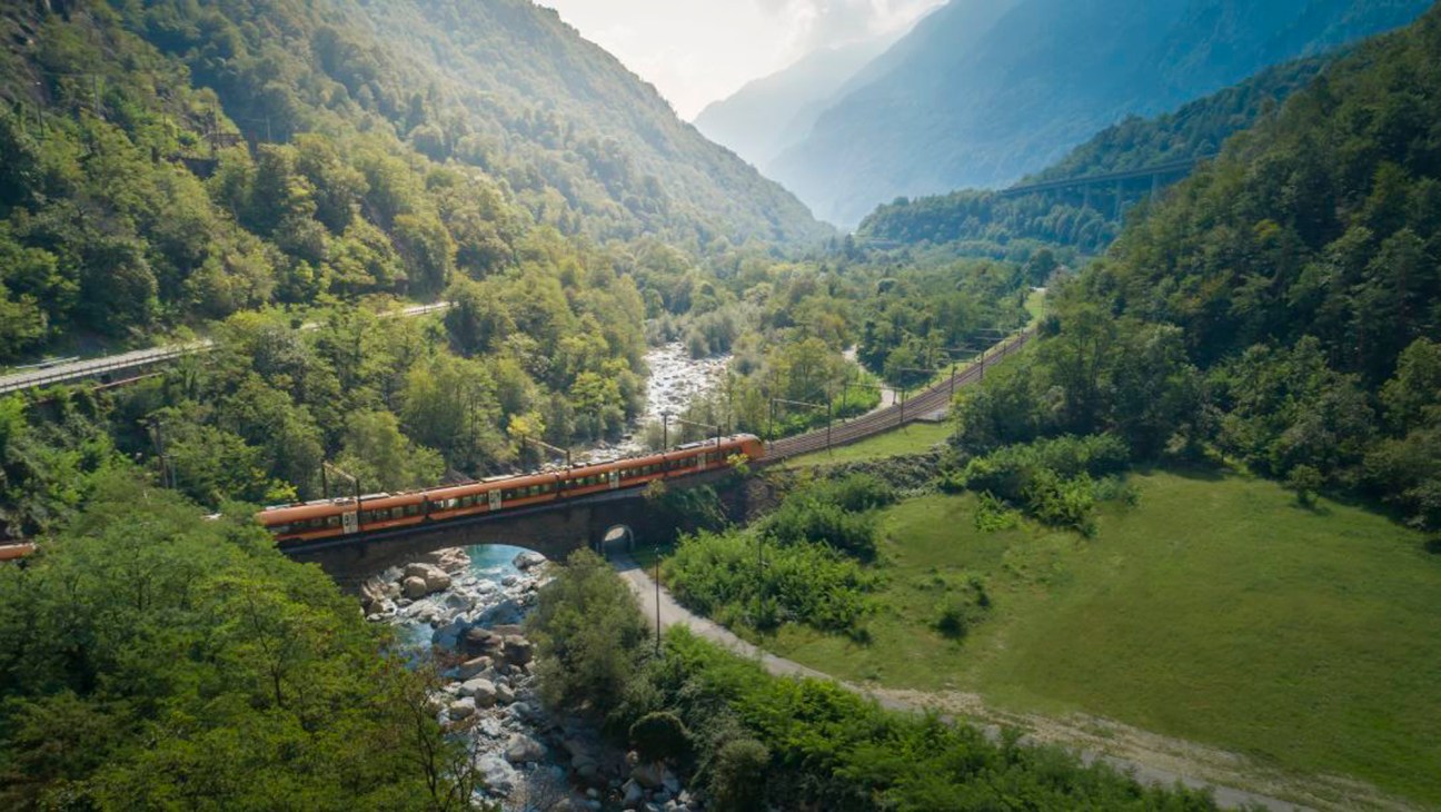 Orange Treno Gottardo train crosses a bridge over a river through a forested Alpine valley in Switzerland.