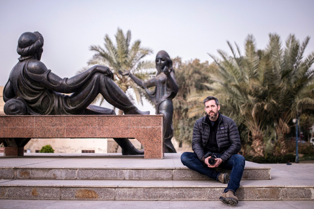 Man with beard in dark jacket sitting on stone steps in front of bronze sculpture and palm trees