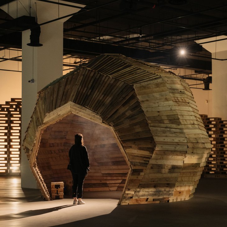 A person stands inside a large wooden tunnel-like installation in an industrial warehouse space.