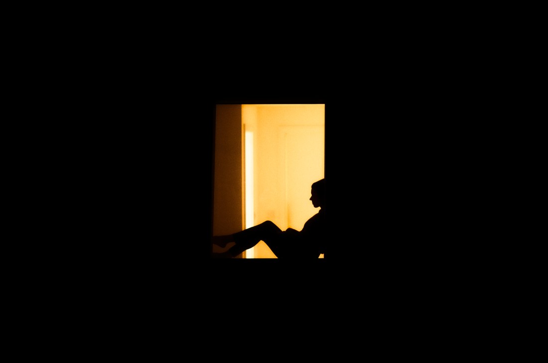 Silhouette of a person leaning against a window with warm golden light streaming through from inside.