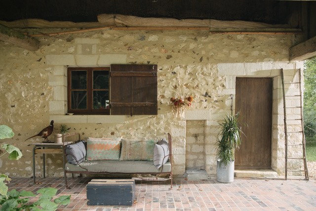 Stone cottage exterior with cushioned seating area, wooden door, and potted plants under shade.