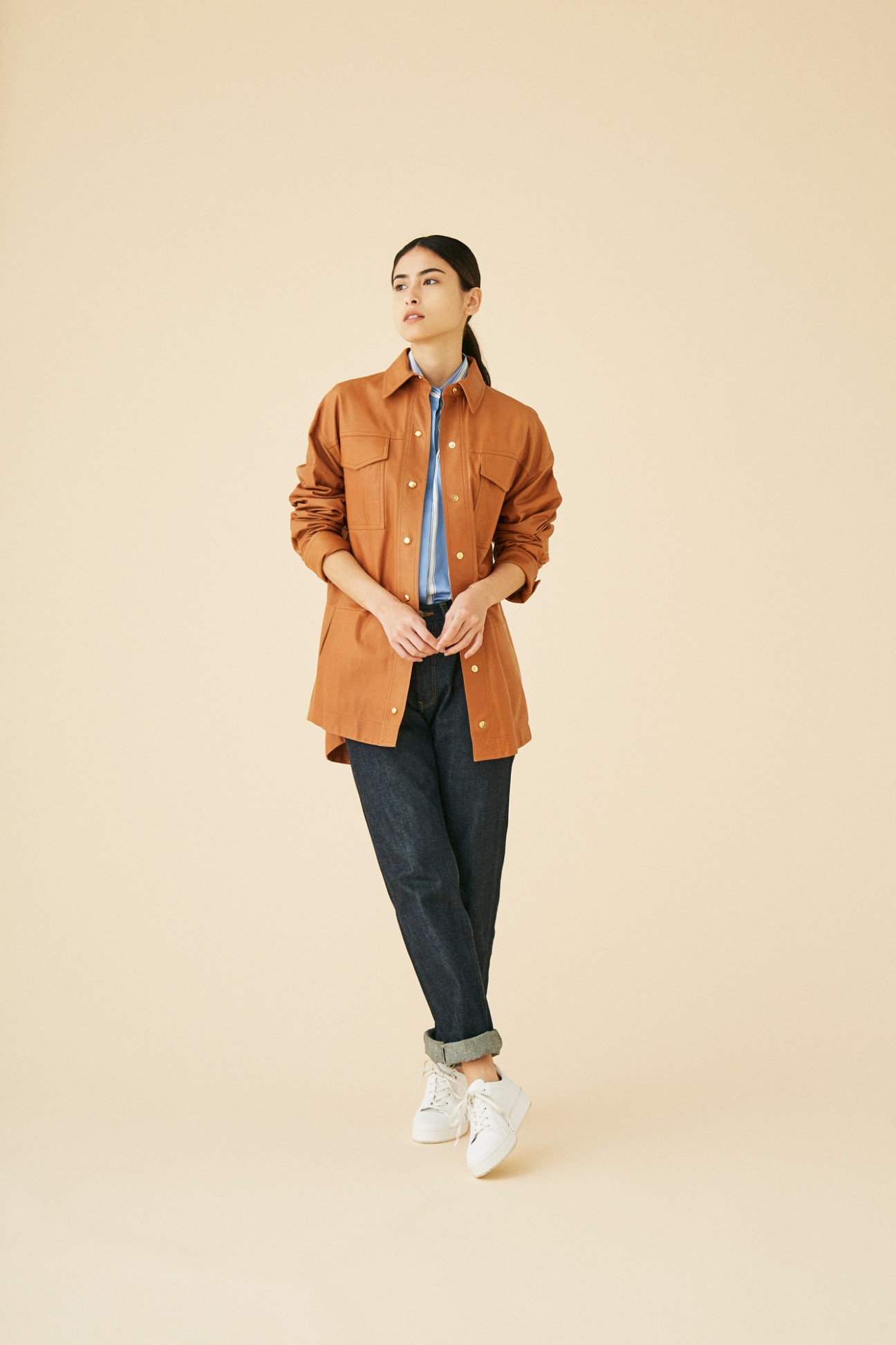 Woman wearing brown shirt jacket over blue shirt with dark jeans and white sneakers against beige background