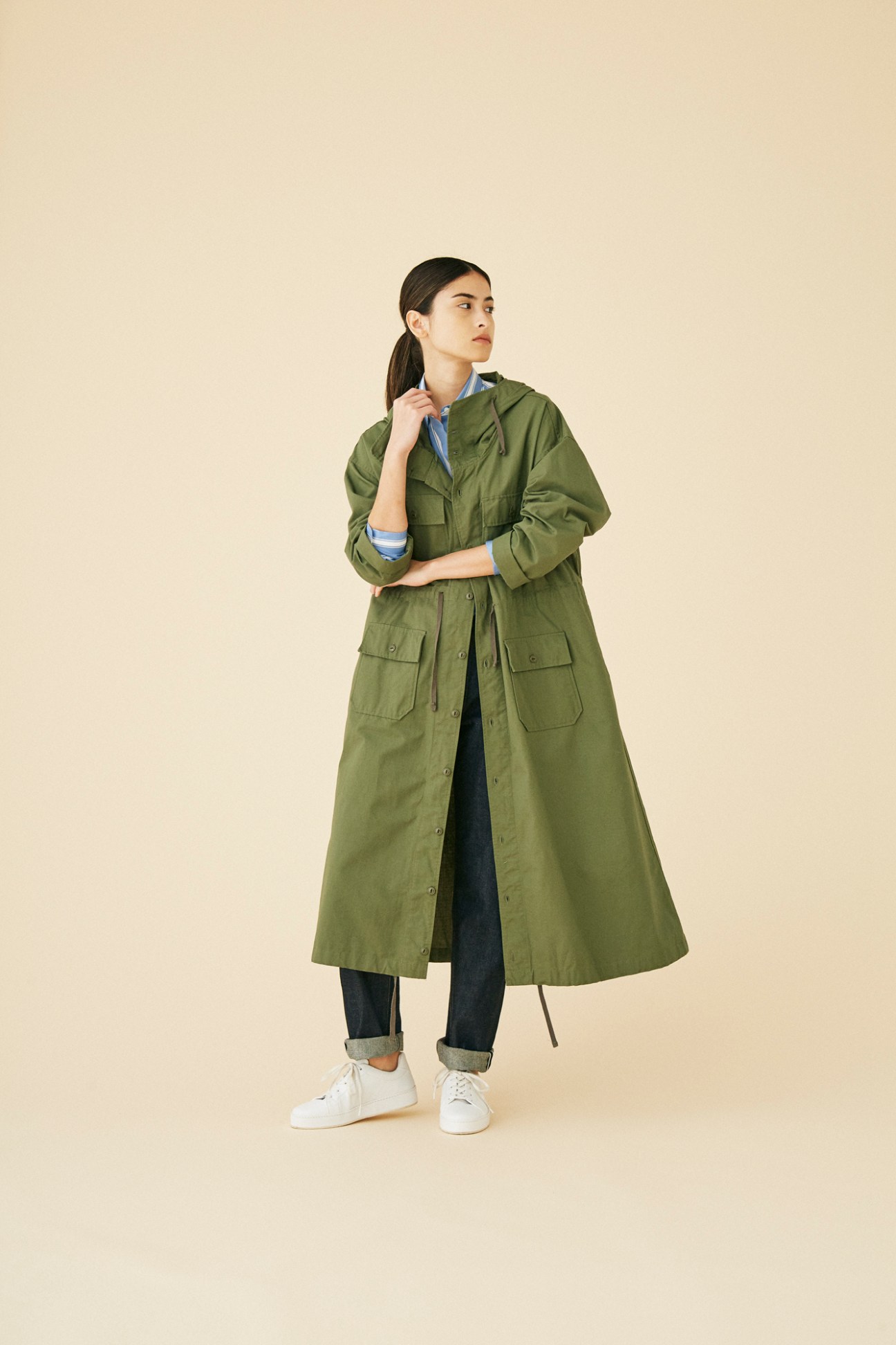 Woman wearing olive green long coat by Engineered Garments with rolled jeans and white sneakers