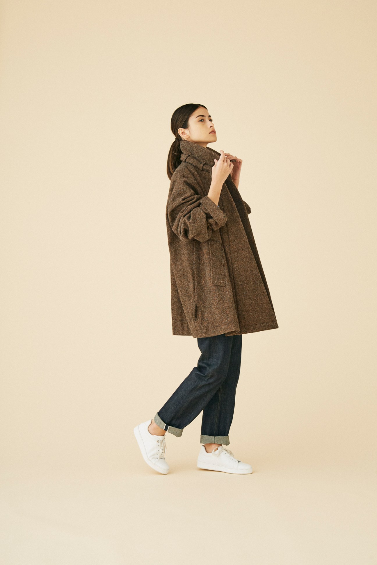 Woman wearing brown wool coat by Lemaire with jeans and white sneakers against beige background
