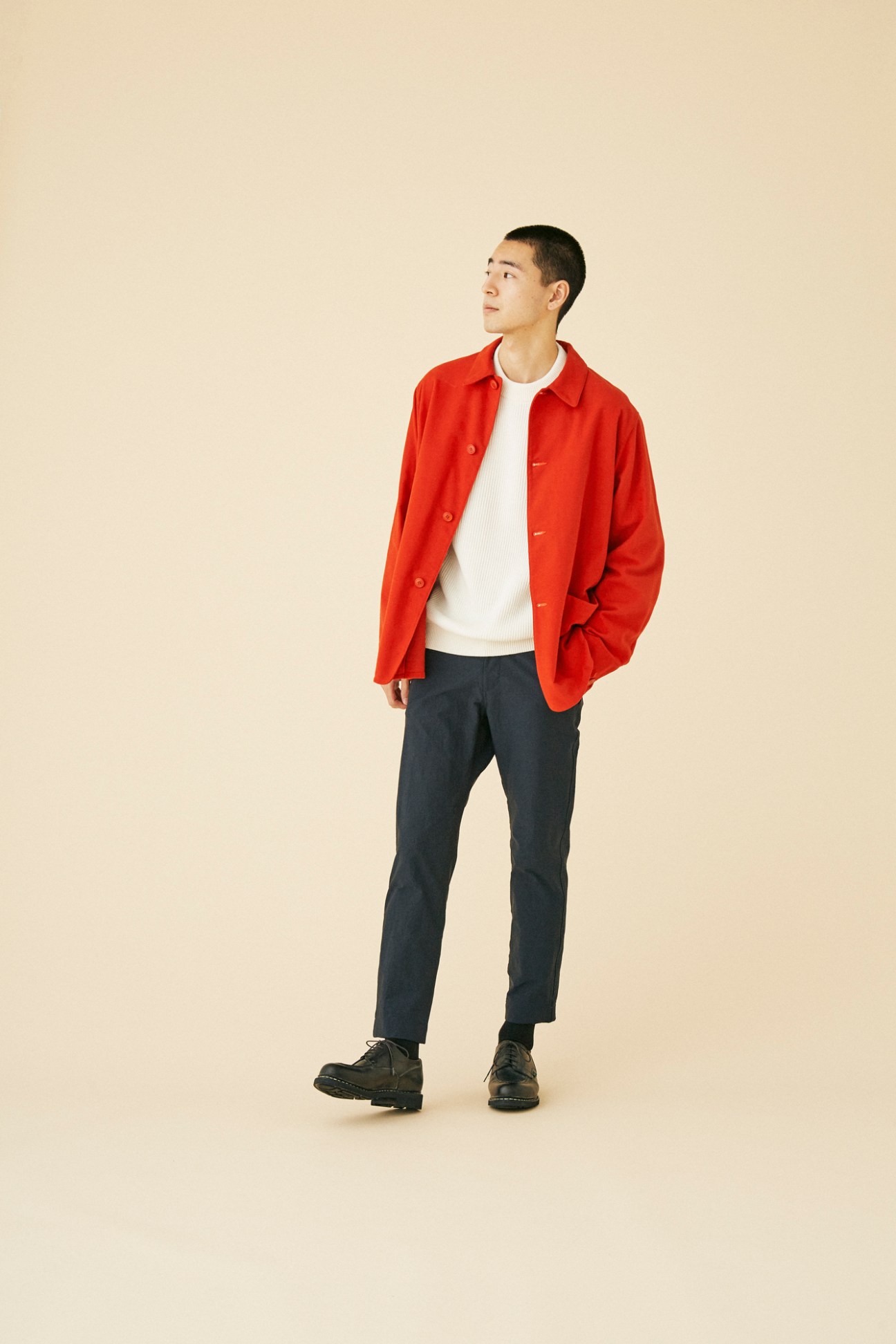 Man wearing red wool jacket by Comoli over white sweater and dark trousers against neutral background