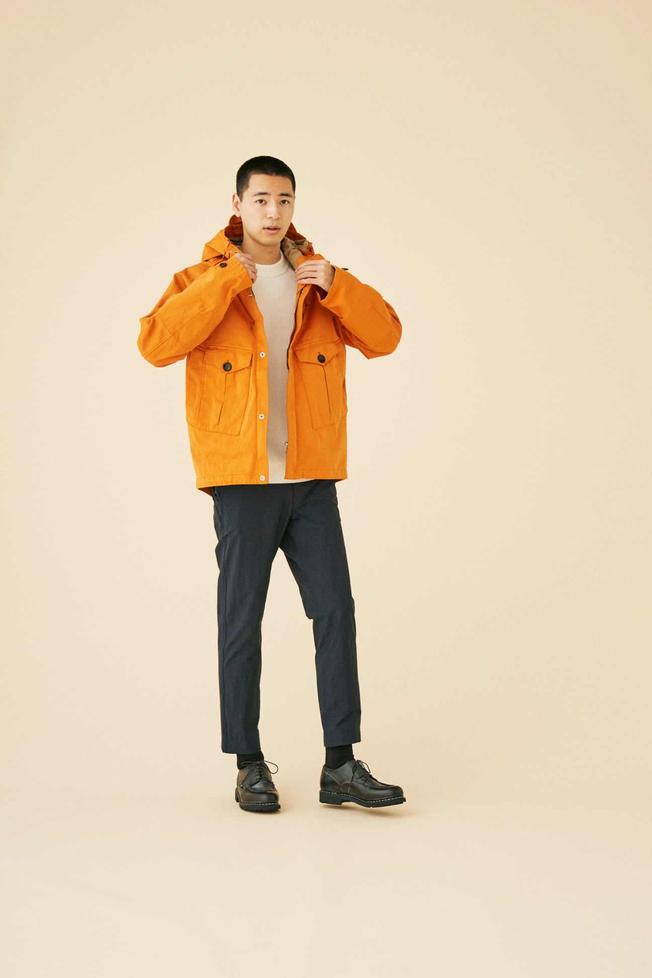 Man wearing bright orange hooded jacket by Ten C with dark trousers and boots against neutral background