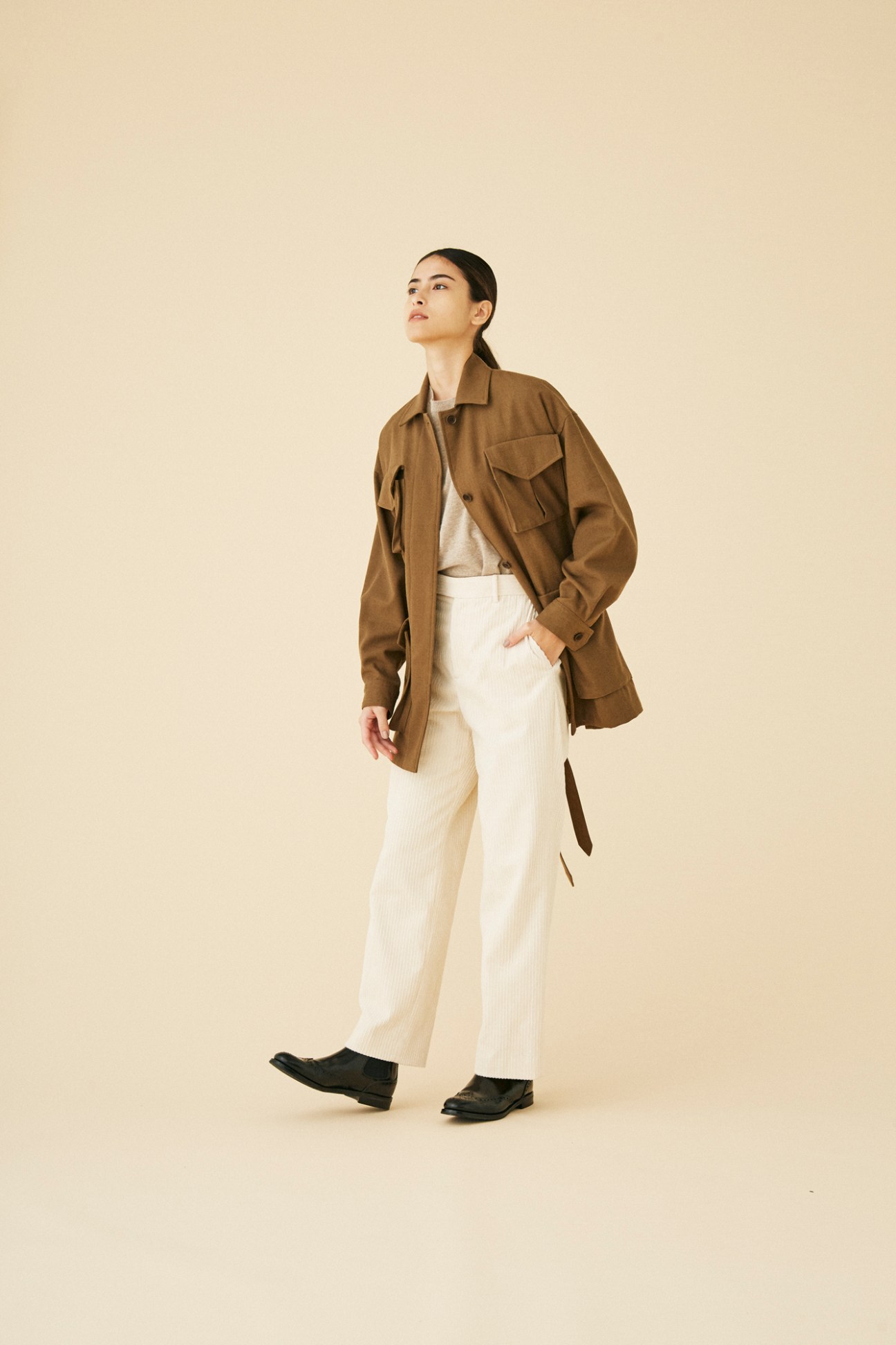Woman wearing brown utility jacket by Blurhms with white trousers in studio setting